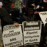 Nighttime street protest with people behind metal barricades holding signs—one reads 'Judaism condemns Zionist Terrorism in Gaza' and another says 'Zionism and Judaism are diametrically opposed'.