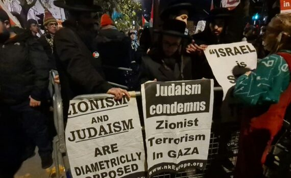 Nighttime street protest with people behind metal barricades holding signs—one reads 'Judaism condemns Zionist Terrorism in Gaza' and another says 'Zionism and Judaism are diametrically opposed'.